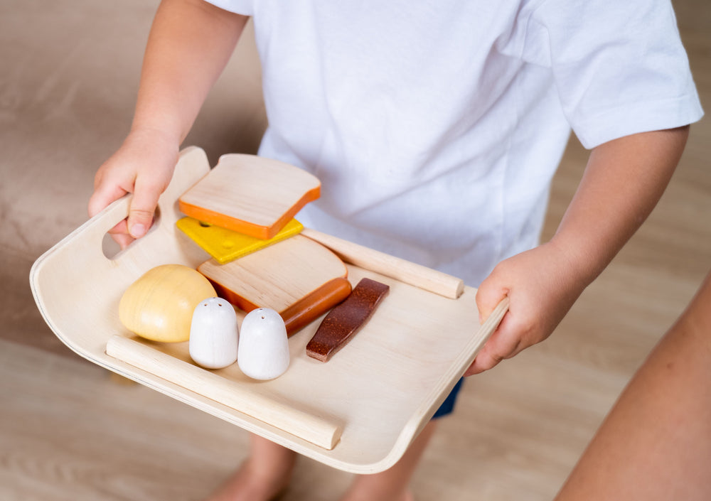 Set Colazione in legno , "Breakfast", Plan Toys. Bambino che tiene in mano vassoio con componenti della colazione.