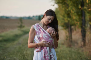 Fascia rigida Dock Dahlia, Huggyberry. Fascia con ordita rosa cipria e rondini bianche stilizzate. Mamma che porta il bambino nella posizione cuore a cuore.