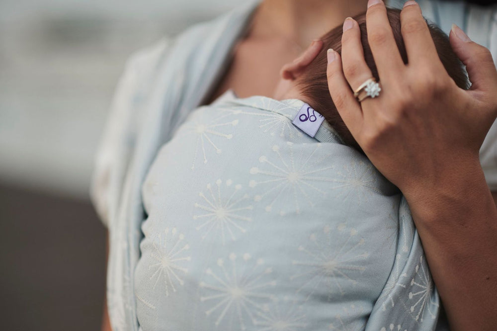 Fascia rigida Dandelion Ice. Mamma che sorregge la testa del suo bambino, legato nella fascia nella posizione cuore a cuore. Particolare dell'ordito color ghiaccio con soffioni bianchi.