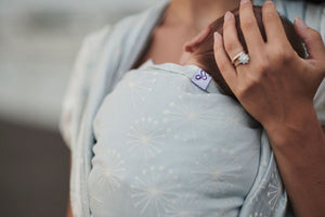 Fascia rigida Dandelion Ice. Mamma che sorregge la testa del suo bambino, legato nella fascia nella posizione cuore a cuore. Particolare dell'ordito color ghiaccio con soffioni bianchi.