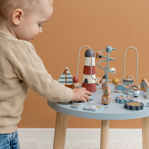 Activity Table, tavolino multiattività in legno, Little Dutch. Tavolino a tema Baia dei Marinai con elementi che richiamano il mare e la pesca. Il tavolino è azzurro con gambe color marrone chiaro. Il gioco impilabile rappresenta un faro, gli ingranaggi richiamano un timone, un salvagente e un gabbiano. Bambino che gioca