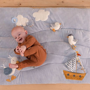 Tappeto morbido PlayPen Mat a tema Baia dei Marinai, Little Dutch. Tappeto rettangolare azzurro, morbido con diverse texture e applicazioni a tema marino tra cui una barchetta a vela con specchietto, uccelli e nuvole. Bambino coricato sul tappeto che sorride
