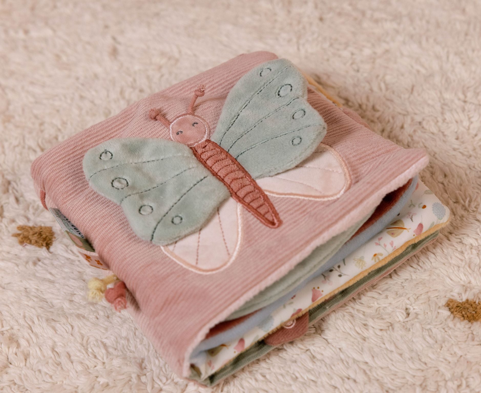 Libro di attività morbido multisensoriale, Little Dutch. Libretto a tema Fiori e Farfalle chiuso e appoggiato sul tappeto. Fronte del libro con farfalla ricamata sopra