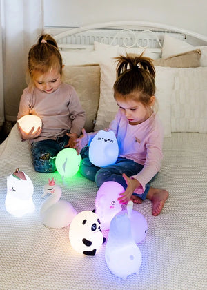 Luce a led notturna in silicone MIDI, InnoGIO. Lampada in silicone bianco a forma di vari animali. Bambine sul letto che giocano con le lampade, alcune accese ed altre spente